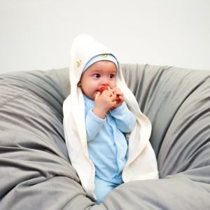En liten bebis sitter på en grå puff. Runt sig har han en beige filt med huva som han har över huvudet.
