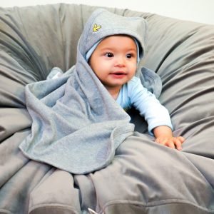 En liten bebis sitter på en grå puff. Runt sig har han en grå filt med huva som han har över huvudet.