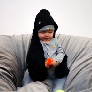 En liten bebis sitter på en grå puff. Runt sig har han en svart filt med huva som han har över huvudet.