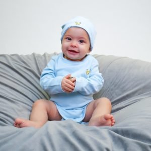 En liten bebis sitter på en stor kudde. På sig har bebisen en ljusblå body med matchande mössa från Lyle & Scott. På bröstet och mössan finns den klassiska gula Lyle & Scott örnen i mindre format